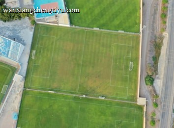 An aerial view of a sports complex featuring multiple green grass fields, including football pitches with marked goalposts. Adjacent to the fields is a blue-roofed building and a smaller area with a tennis or basketball court. Surrounding the complex, there's a road lined with trees and a dirt area with sparse vegetation.